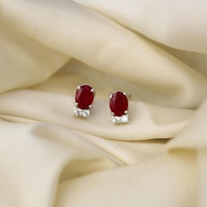 以下が含まれることがあります： 楕円形の赤い宝石が、より小さな透明な宝石で囲まれた、銀製のスタッドピアスのペアです。