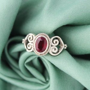 Può includere: Un anello d'argento con una pietra preziosa rossa incastonata in un design a spirale decorativo.