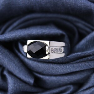 Puede incluir: Un anillo de plata con una piedra ovalada negra y pequeñas piedras transparentes en la banda.
