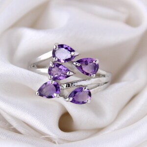 Peut inclure: Une bague en argent avec cinq pierres précieuses améthystes en forme de goutte. La bague est posée sur un fond de tissu blanc.