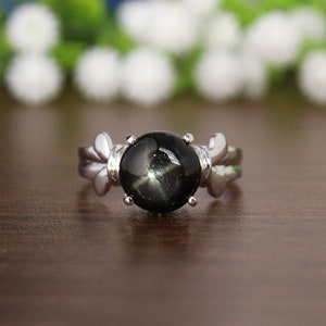 Könnte beinhalten: Ein silberner Ring mit einem schwarzen sternförmigen Edelstein. Der Ring hat ein zartes Design mit zwei kleinen Flügeln auf jeder Seite des Steins.
