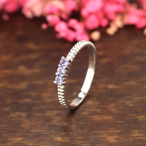 Peut inclure: Une bague en argent avec trois petites pierres précieuses violettes serties en rangée. La bague a une bande texturée.