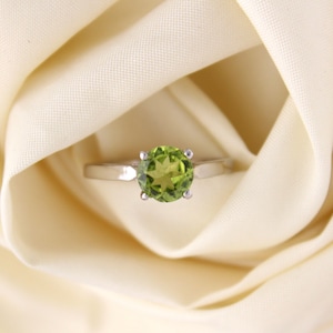 Peut inclure: Une bague en argent avec une pierre précieuse verte sertie à quatre griffes. La bague est sur un fond de tissu blanc.