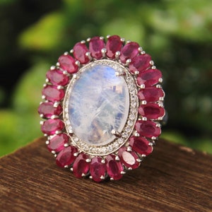 May include: A silver ring featuring a large oval moonstone set within a halo of small red gemstones.