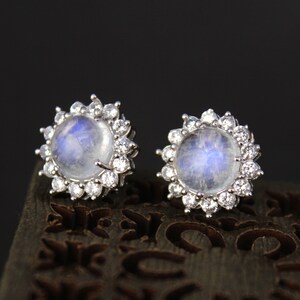 Può includere: Un paio di orecchini d'argento con un centro rotondo di pietra di luna circondato da un alone di pietre più piccole e trasparenti.