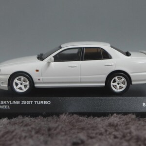 Könnte beinhalten: Ein weißes Nissan Skyline 25GT Turbo Nismo Wheel Modellauto auf einem schwarzen Präsentationsständer. Das Modellauto hat weiße Räder und einen Heckspoiler. Der Text auf dem Ständer lautet "NISSAN SKYLINE 25GT TURBO NISMO WHEEL" und "816pcs."