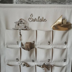 Countdown to Christmas with this canvas advent calendar which can be personalized with name. Featuring 24 pockets to mark each day of December, it can be filled with small gifts. Off white, sand, ecru colors. Handmade product. With wooden numbers.