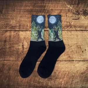 May include: A pair of black socks with a forest and full moon design. The design features a silhouette of a creature reaching up to the moon.