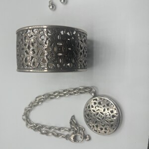 May include: A silver-toned jewelry set featuring a cuff bracelet, a pendant necklace, and a pair of stud earrings. The bracelet and pendant have an openwork design. The necklace has a chain with a lobster clasp.