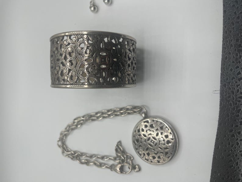 May include: A silver-toned jewelry set featuring a cuff bracelet, a pendant necklace, and a pair of stud earrings. The bracelet and pendant have an openwork design. The necklace has a chain with a lobster clasp.