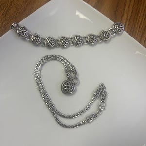 Puede incluir: Dos pulseras plateadas sobre una superficie blanca. Una presenta eslabones en forma de coraz&oacute;n, la otra es una cadena con un dije de coraz&oacute;n. Ambas probablemente de metal.