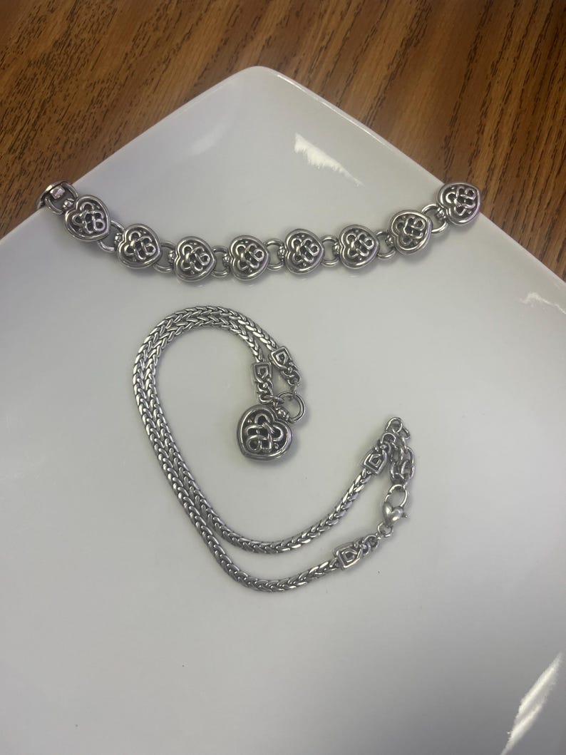 Puede incluir: Dos pulseras plateadas sobre una superficie blanca. Una presenta eslabones en forma de coraz&oacute;n, la otra es una cadena con un dije de coraz&oacute;n. Ambas probablemente de metal.