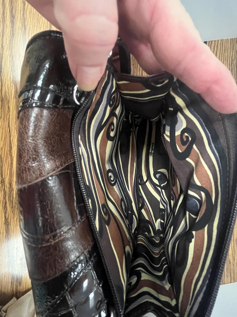 May include: A brown leather handbag with a glossy finish. The bag features a patchwork design with varying shades of brown and a patterned interior lining. The zipper is open, revealing the inside of the bag.