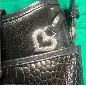 May include: Close-up of a black leather handbag with a silver heart-shaped charm. The charm is engraved with the brand name "Brighton". The bag has a textured, crocodile-like pattern on the lower portion.
