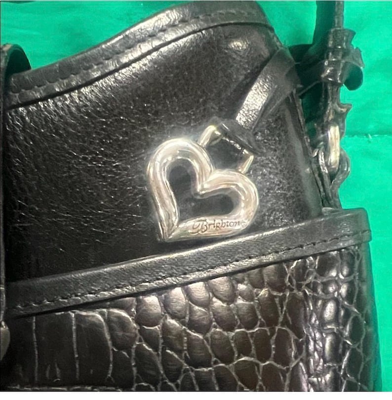 May include: Close-up of a black leather handbag with a silver heart-shaped charm. The charm is engraved with the brand name "Brighton". The bag has a textured, crocodile-like pattern on the lower portion.