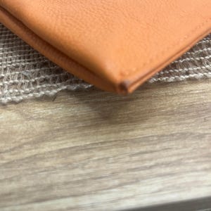 May include: A folded orange leather wallet featuring a smooth, grained texture and visible stitched edges along the perimeter. The material displays a consistent vibrant orange hue with a slight darkening at the corner fold of the leather item.