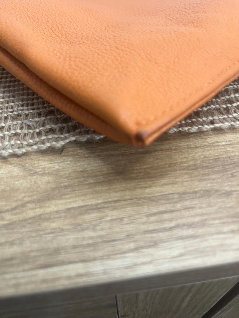 May include: A folded orange leather wallet featuring a smooth, grained texture and visible stitched edges along the perimeter. The material displays a consistent vibrant orange hue with a slight darkening at the corner fold of the leather item.