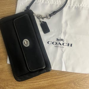 May include: A black leather wristlet featuring a silver-toned turn-lock closure on the front flap, a detachable wrist strap, and a small leather hangtag. The wristlet is displayed next to a white fabric dust bag printed with the Coach New York logo.