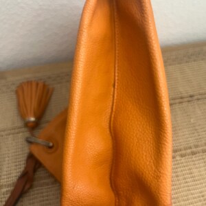 May include: A bright orange leather clutch bag with a textured surface. The bag features a silver-toned metal ring attachment connected to a braided leather strap and a matching orange leather tassel hanging from the side of the main body.