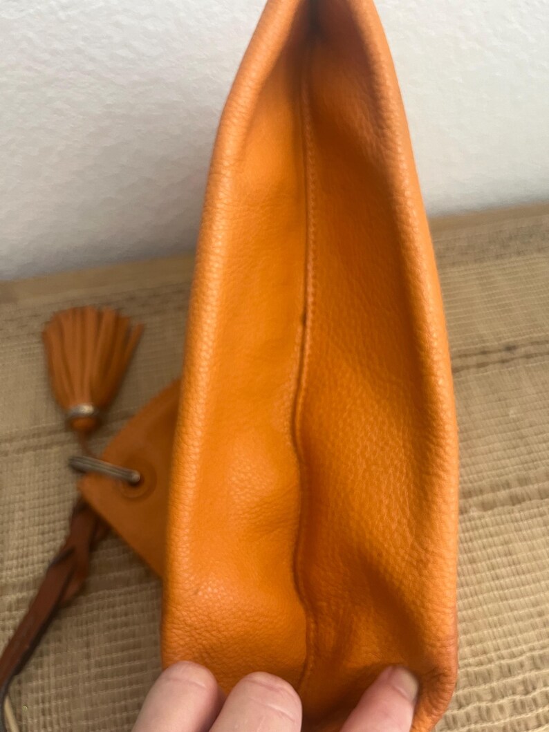 May include: A bright orange leather clutch bag with a textured surface. The bag features a silver-toned metal ring attachment connected to a braided leather strap and a matching orange leather tassel hanging from the side of the main body.