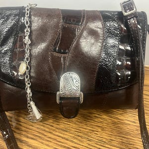 May include: A brown and black leather crossbody bag with a silver clasp and strap. The bag has a patchwork design with a shiny, textured finish. A silver chain with charms hangs from the top.