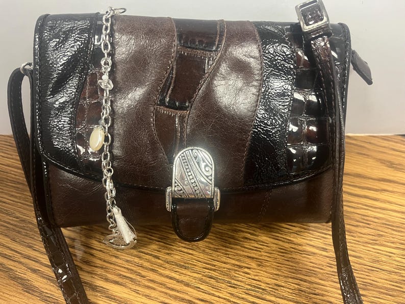 May include: A brown and black leather crossbody bag with a silver clasp and strap. The bag has a patchwork design with a shiny, textured finish. A silver chain with charms hangs from the top.