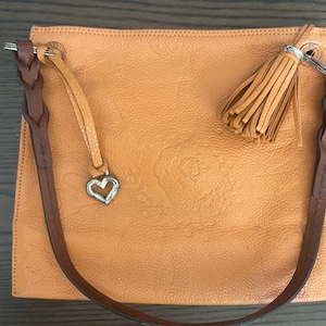 May include: A square-shaped orange handbag with a textured surface, featuring a dark brown braided shoulder strap, a matching orange leather tassel, and a silver-toned heart-shaped charm attached to a thin leather cord on the top left corner.