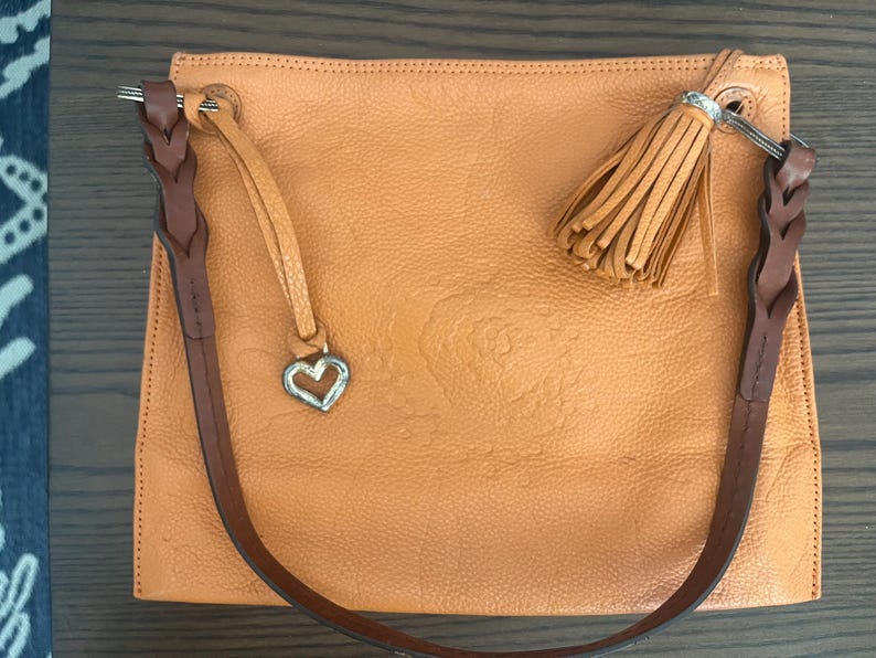 May include: A square-shaped orange handbag with a textured surface, featuring a dark brown braided shoulder strap, a matching orange leather tassel, and a silver-toned heart-shaped charm attached to a thin leather cord on the top left corner.