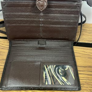 May include: Open brown leather wallet with multiple card slots and a clear ID window. The wallet has a heart-shaped clasp and a zipped compartment. The interior is a lighter shade of brown than the exterior. The wallet is open, revealing its contents.