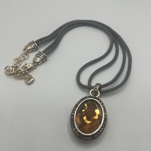 May include: A silver-toned necklace with a gray cord. The pendant is oval-shaped with a brown center and a silver border with small, clear stones. The necklace has a lobster clasp.