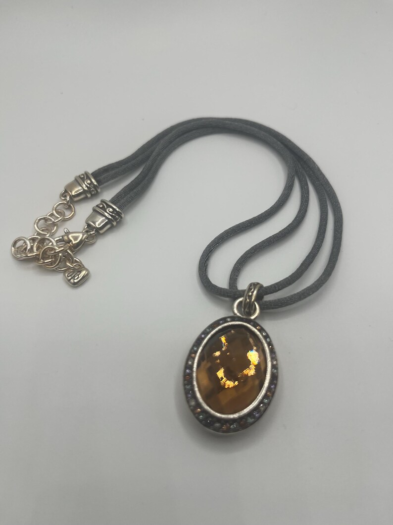May include: A silver-toned necklace with a gray cord. The pendant is oval-shaped with a brown center and a silver border with small, clear stones. The necklace has a lobster clasp.
