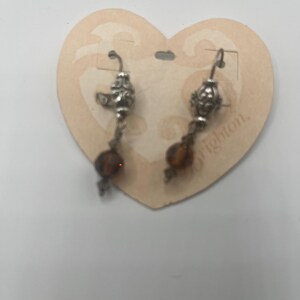 May include: Silver-toned dangle earrings with ornate filigree details and amber-colored beads. The earrings are displayed on a heart-shaped cardboard tag. The word "Brighton" is partially visible on the tag.
