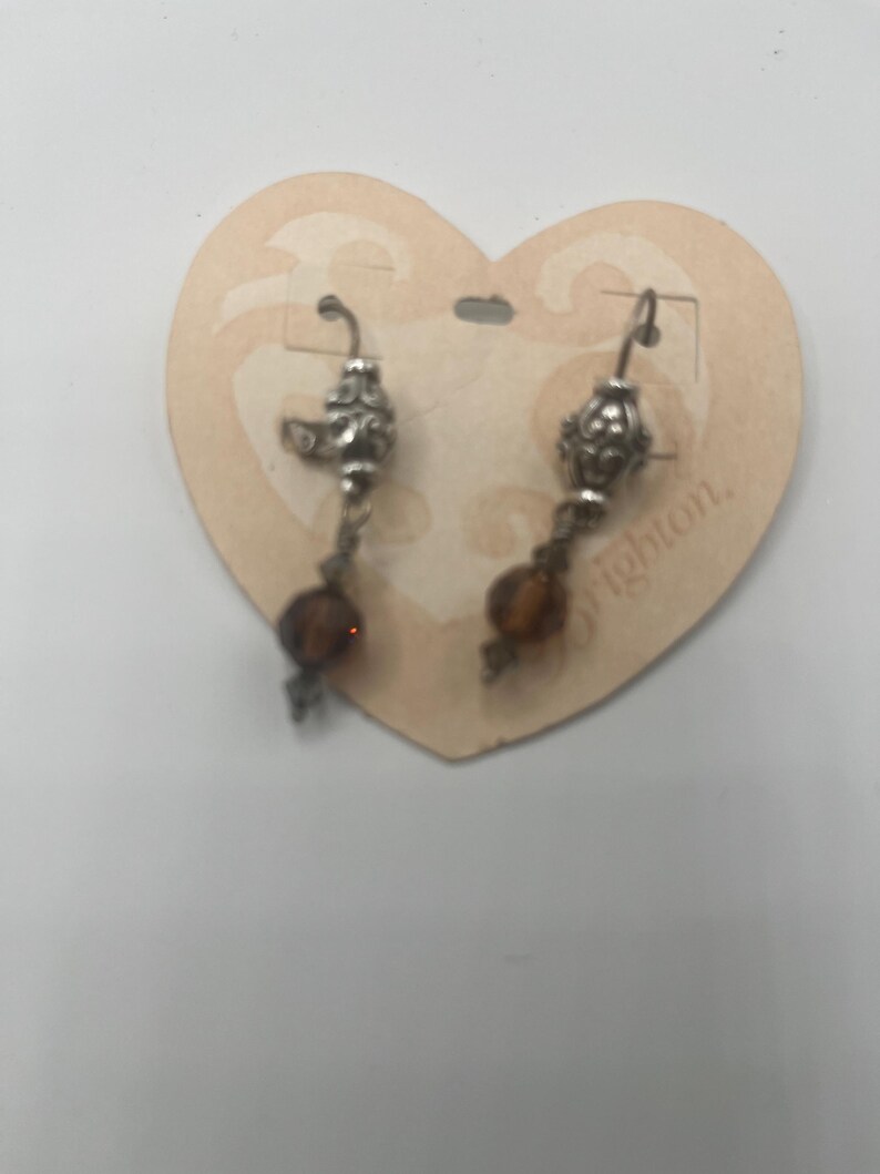 May include: Silver-toned dangle earrings with ornate filigree details and amber-colored beads. The earrings are displayed on a heart-shaped cardboard tag. The word "Brighton" is partially visible on the tag.