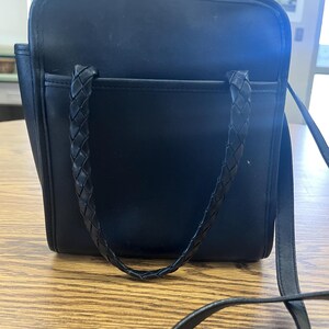 May include: Black square handbag with a braided handle and a long shoulder strap. The bag has a front pocket and is sitting on a wooden surface. The bag is made of a smooth material.