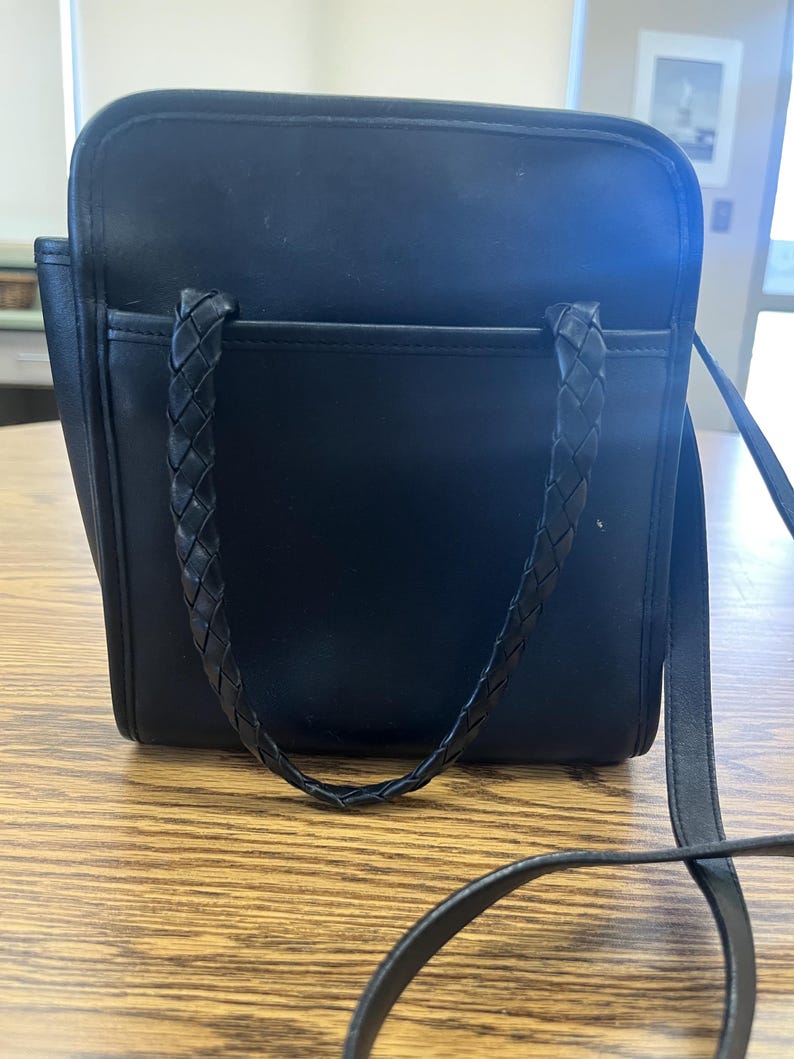 May include: Black square handbag with a braided handle and a long shoulder strap. The bag has a front pocket and is sitting on a wooden surface. The bag is made of a smooth material.