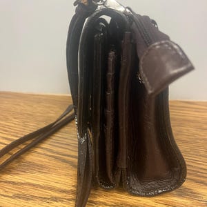 May include: A dark brown leather crossbody bag with a long strap. The bag has a rectangular shape with a flap closure and a zipper pocket. The leather has a glossy finish and a silver-coloured clasp.