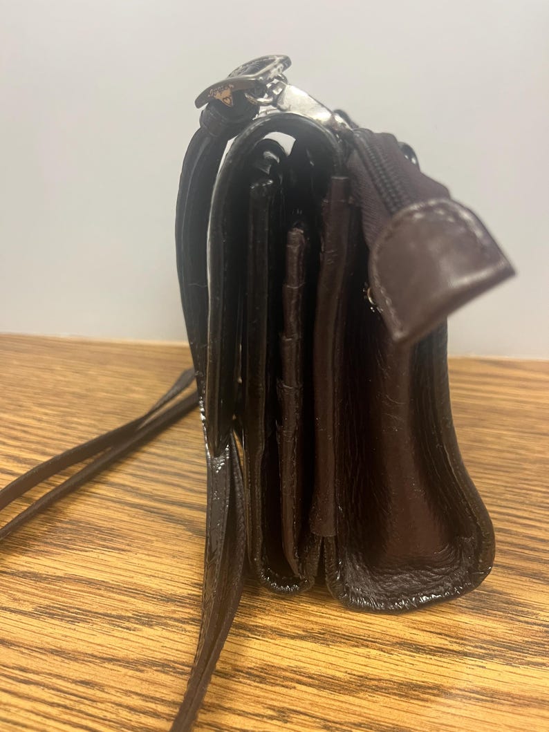 May include: A dark brown leather crossbody bag with a long strap. The bag has a rectangular shape with a flap closure and a zipper pocket. The leather has a glossy finish and a silver-coloured clasp.