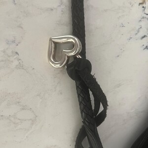 Puede incluir: Primer plano de una correa de bolso de cuero negro con un dije plateado en forma de coraz&oacute;n. La correa est&aacute; trenzada y sujeta a la bolsa con un detalle de cuero anudado. El fondo es una superficie clara y moteada.