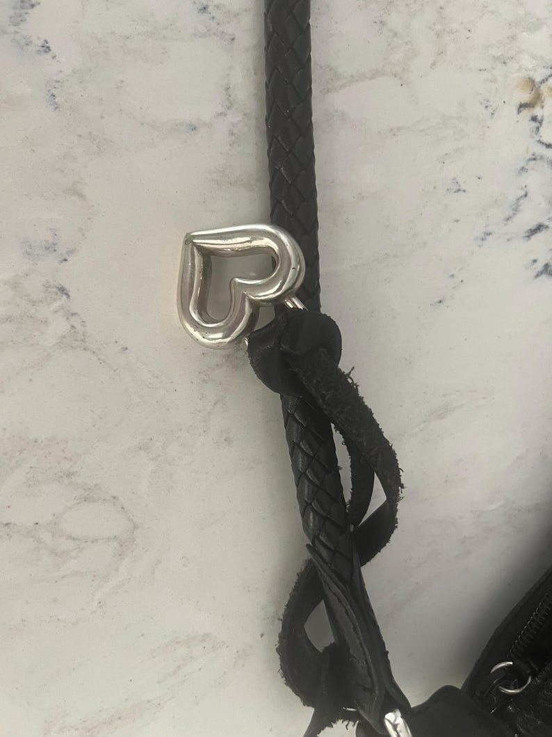 Puede incluir: Primer plano de una correa de bolso de cuero negro con un dije plateado en forma de coraz&oacute;n. La correa est&aacute; trenzada y sujeta a la bolsa con un detalle de cuero anudado. El fondo es una superficie clara y moteada.