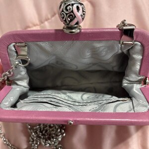 May include: A pink leather purse with a silver chain strap. The purse has a silver clasp and a decorative top with a pink ribbon design. The interior is lined with a gray patterned fabric.