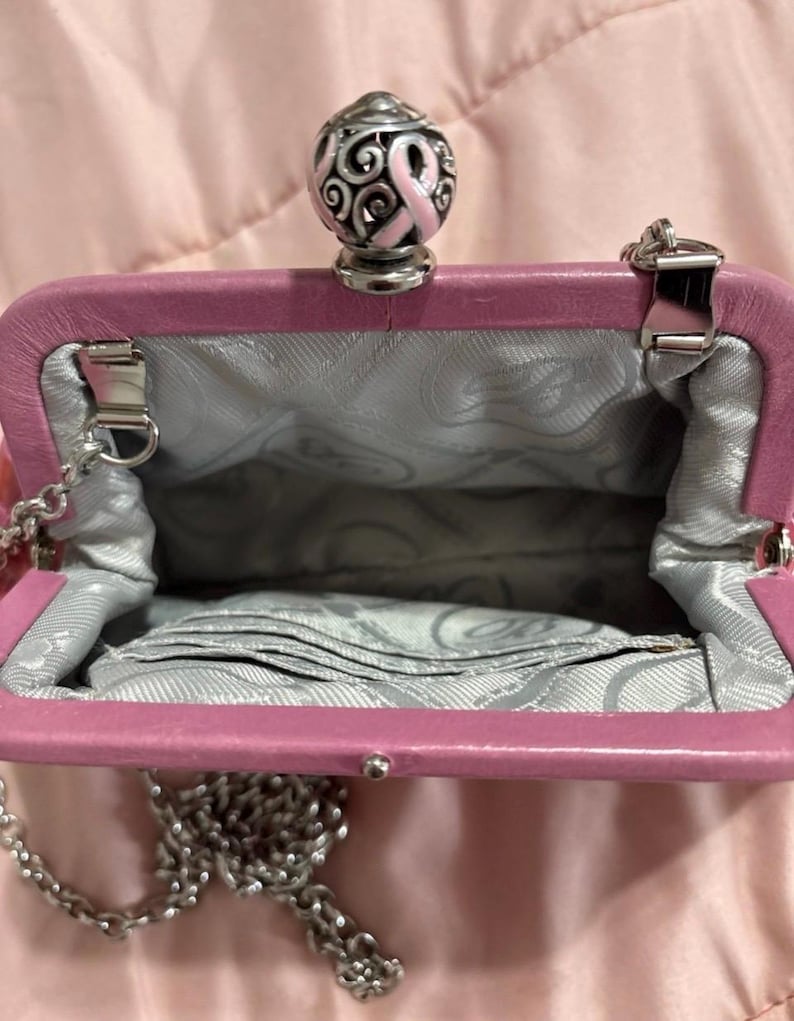 May include: A pink leather purse with a silver chain strap. The purse has a silver clasp and a decorative top with a pink ribbon design. The interior is lined with a gray patterned fabric.