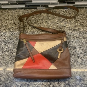 May include: A Fossil crossbody bag featuring a multi-colored geometric leather patchwork design in shades of red, black, metallic gold, and brown. The bag includes a top zipper closure, a long adjustable brown leather strap, and a hanging key charm.