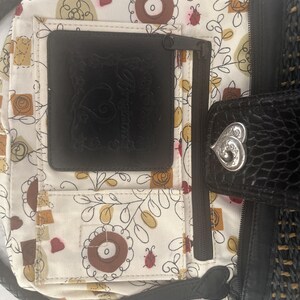 Puede incluir: Bolso de mano negro y tejido con una correa trenzada. El interior presenta un estampado floral con detalles en marr&oacute;n, amarillo y rojo. Se puede ver un bolsillo de cuero negro, junto con un cierre plateado en forma de coraz&oacute;n.