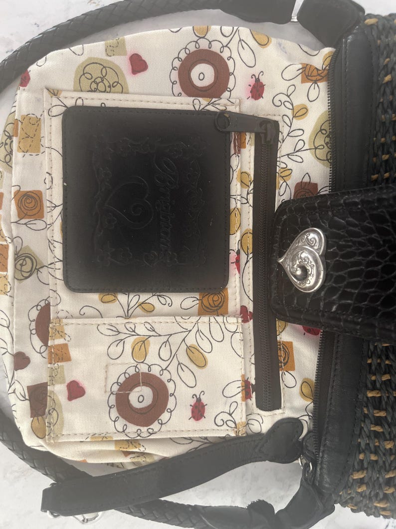 Puede incluir: Bolso de mano negro y tejido con una correa trenzada. El interior presenta un estampado floral con detalles en marr&oacute;n, amarillo y rojo. Se puede ver un bolsillo de cuero negro, junto con un cierre plateado en forma de coraz&oacute;n.