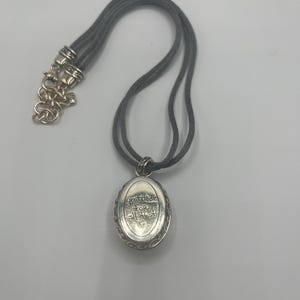 May include: A silver-toned pendant necklace with a gray cord. The oval pendant has the words "gratitude for all that is" engraved on it. The necklace has a decorative clasp.