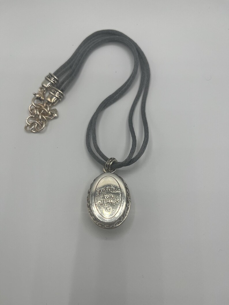 May include: A silver-toned pendant necklace with a gray cord. The oval pendant has the words "gratitude for all that is" engraved on it. The necklace has a decorative clasp.