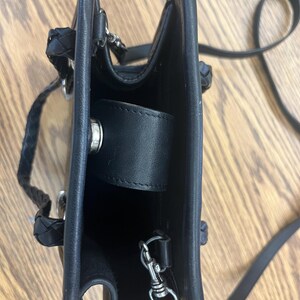 May include: Black leather handbag with a braided handle and a long, thin shoulder strap. The bag has a rectangular shape with a flap closure and silver-colored hardware. The interior is visible.