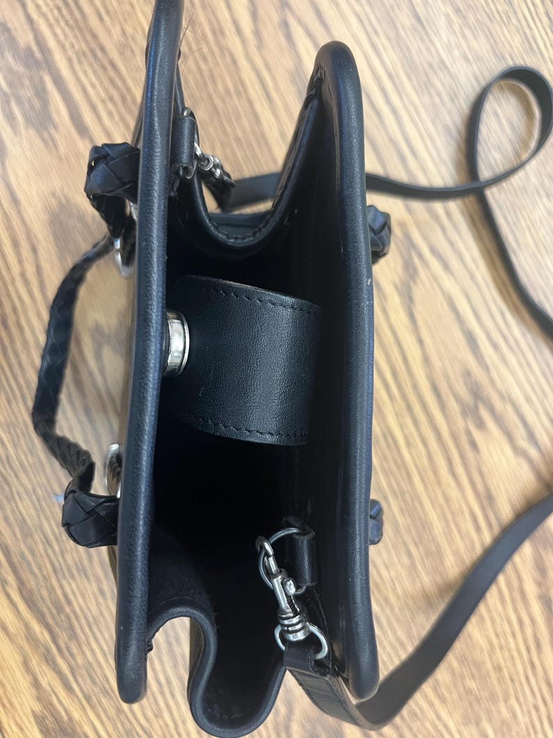 May include: Black leather handbag with a braided handle and a long, thin shoulder strap. The bag has a rectangular shape with a flap closure and silver-colored hardware. The interior is visible.