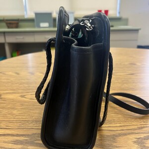 May include: Black leather crossbody bag with a braided strap. The bag has a rectangular shape with rounded corners and a silver zipper. The interior is visible, revealing a glimpse of the contents. The bag is sitting on a wooden table.