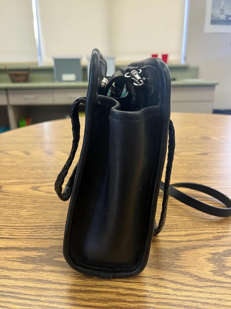 May include: Black leather crossbody bag with a braided strap. The bag has a rectangular shape with rounded corners and a silver zipper. The interior is visible, revealing a glimpse of the contents. The bag is sitting on a wooden table.